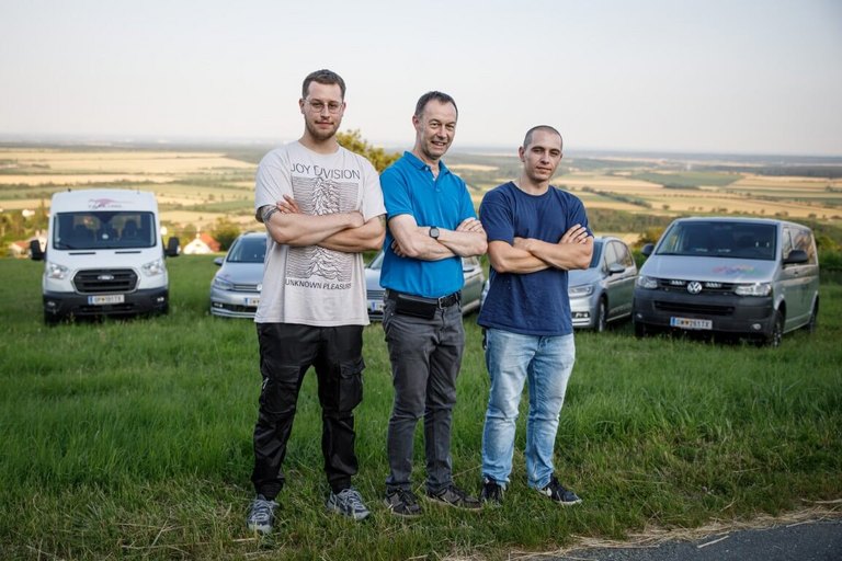 Drei Männer stehen mit verschränkten Armen auf einer Wiese vor mehreren Shuttlefahrzeugen, mit weitem Blick über die Landschaft des Südburgenlands – repräsentiert das Team des Taxi- und Bikeshuttle-Service Loibl in Rechnitz.