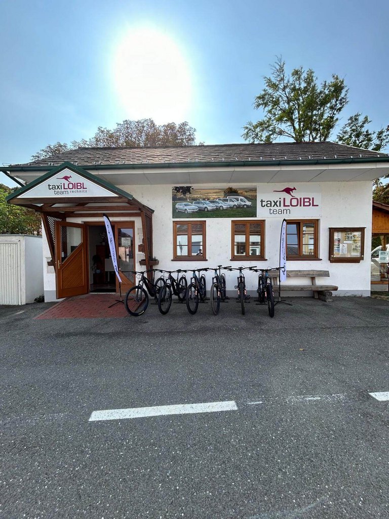 Gebäude des Taxi Loibl Teams in Rechnitz bei Sonnenschein, mit mehreren Mountainbikes zur Miete vor dem Eingang und zwei Fahnen – zentraler Standort für Radverleih, Shuttle-Service und persönliche Beratung.