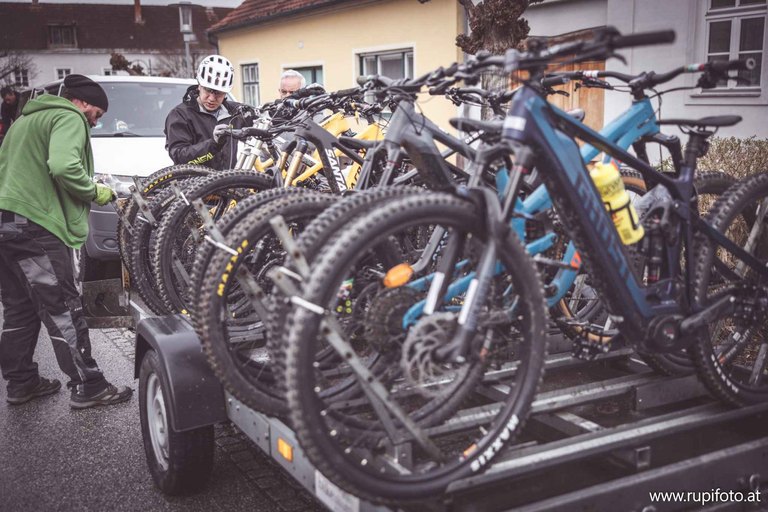 Mehrere hochwertige Mountainbikes auf einem Fahrradanhänger, bereit für den Transport, im Vordergrund ein E-Mountainbike mit Trinkflasche; im Hintergrund zwei Personen beim Beladen vor einem Haus – Teil des Bike-Shuttle-Service rund um den Geschriebenstein.
