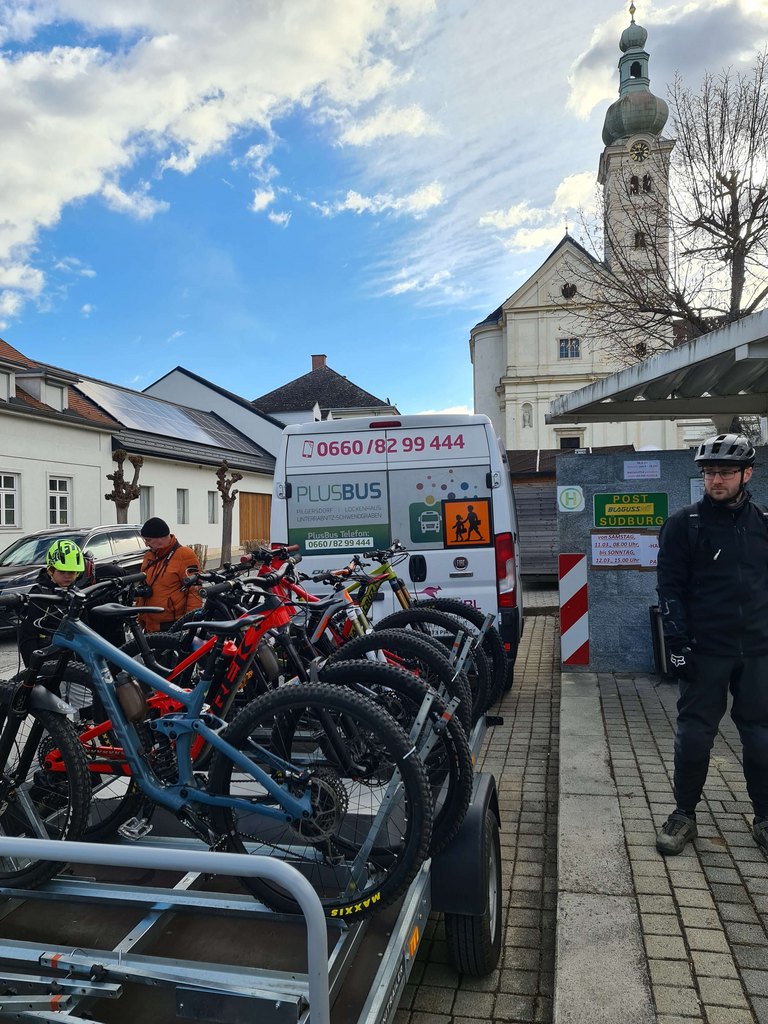 Mehrere moderne Mountainbikes auf einem Anhänger vor einem Shuttlebus mit Aufschrift „PLUSBUS“, geparkt an einer Haltestelle in der Nähe einer Kirche; im Bild auch zwei Radfahrer in Ausrüstung – zeigt den Biketransport-Service im Raum Südburgenland.