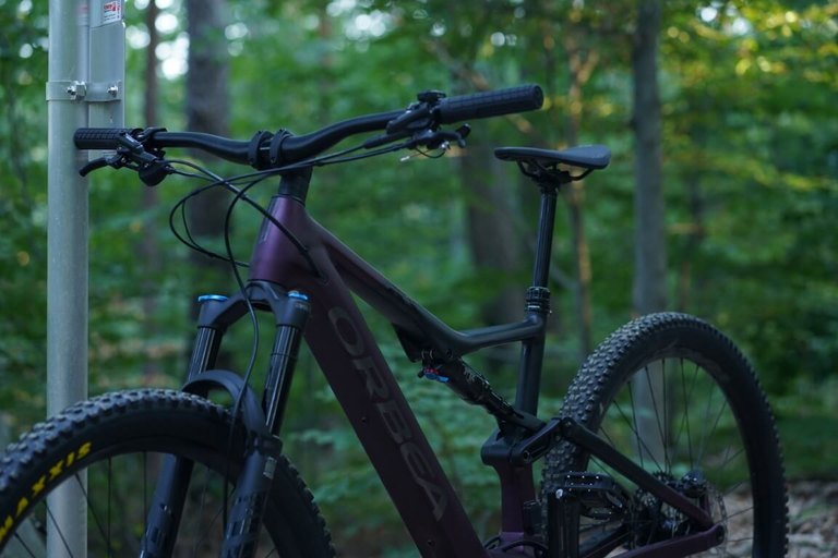 Modernes Orbea Fully-Mountainbike in dunklem Violett, abgestellt im Wald an einem Schildmast, mit breiten Reifen, Federgabel und absenkbarer Sattelstütze – bereit für anspruchsvolle Trails rund um den Geschriebenstein.