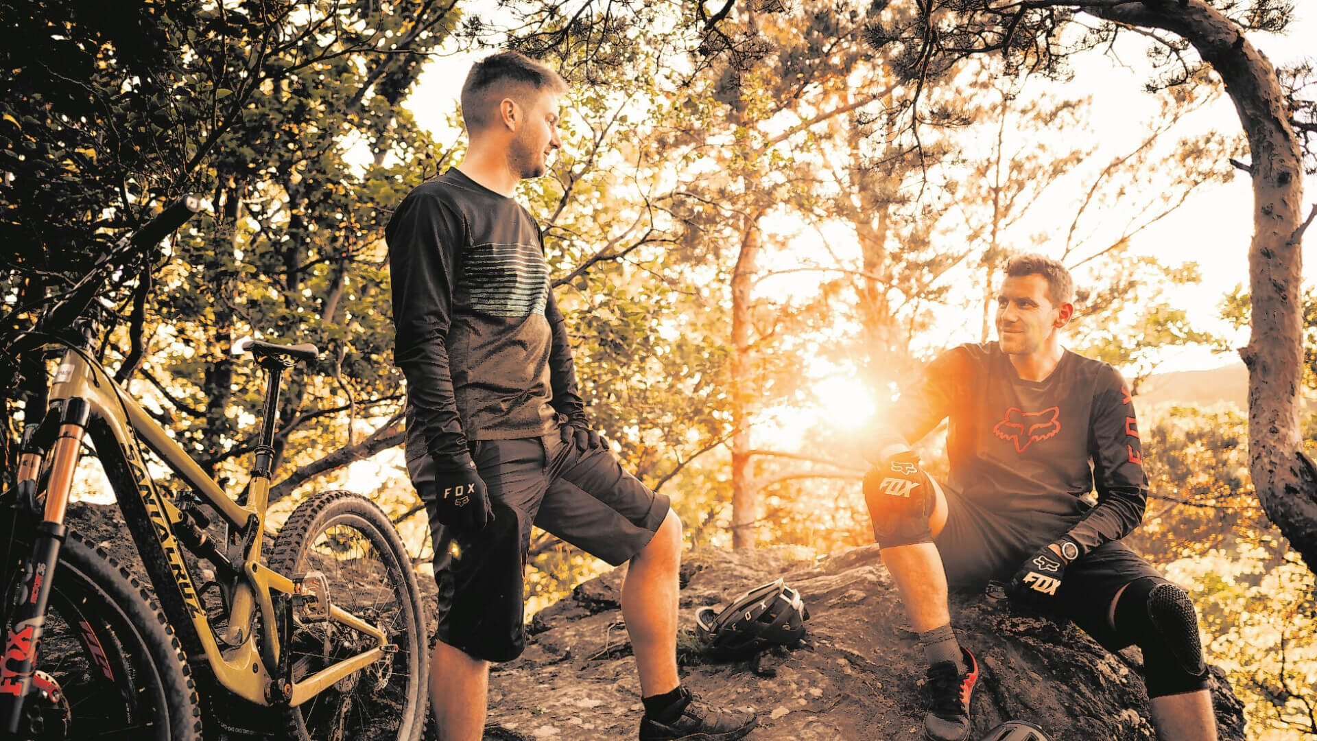 Zwei Mountainbiker in Schutzkleidung machen eine Pause im Wald bei Sonnenuntergang, mit einem Santa Cruz Fahrrad im Vordergrund.